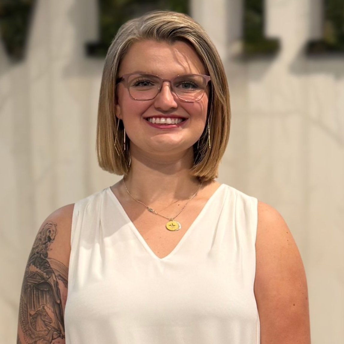 Woman with glasses and blonde bob smiles, wearing a white top and gold necklace.