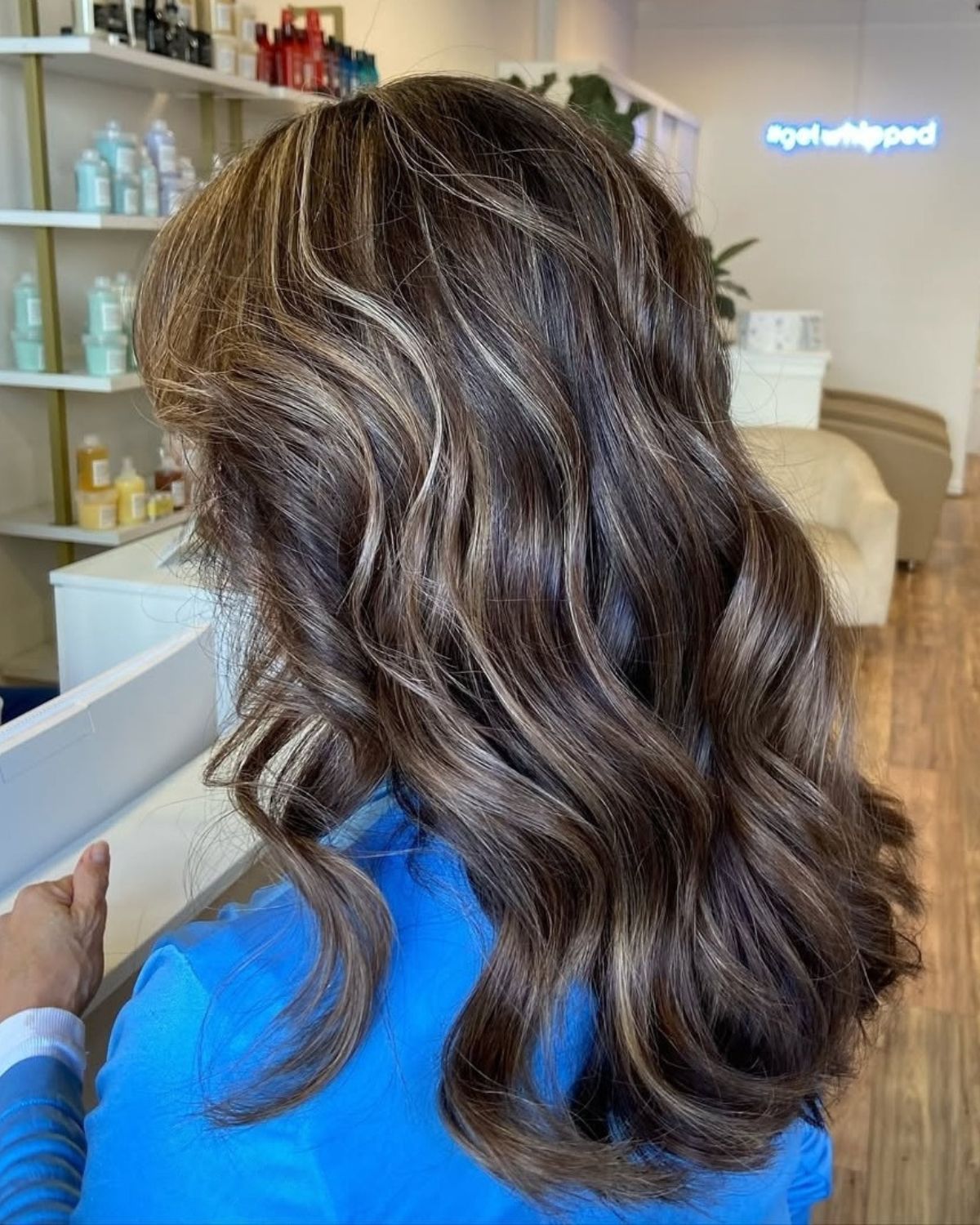 Woman with dark brown wavy hair, highlighted with light brown strands. She is wearing a blue top.