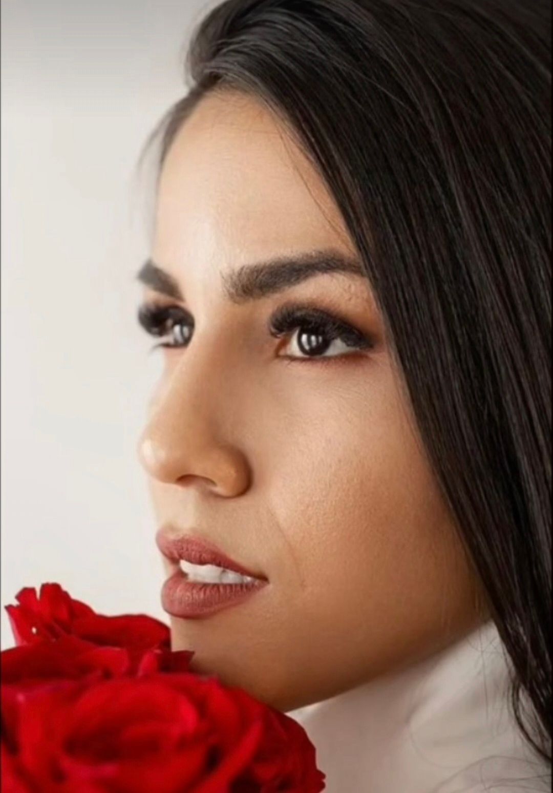 Woman with dark hair, looking to the side, holding red roses, white background.