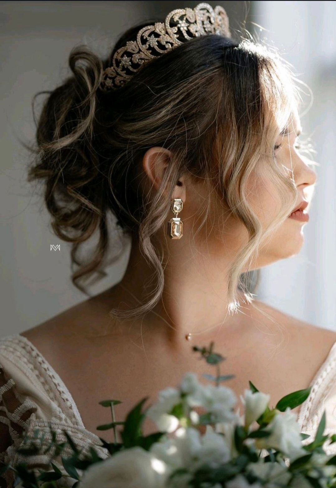 Woman wearing a crown and earrings, looking out a window; bouquet in foreground.