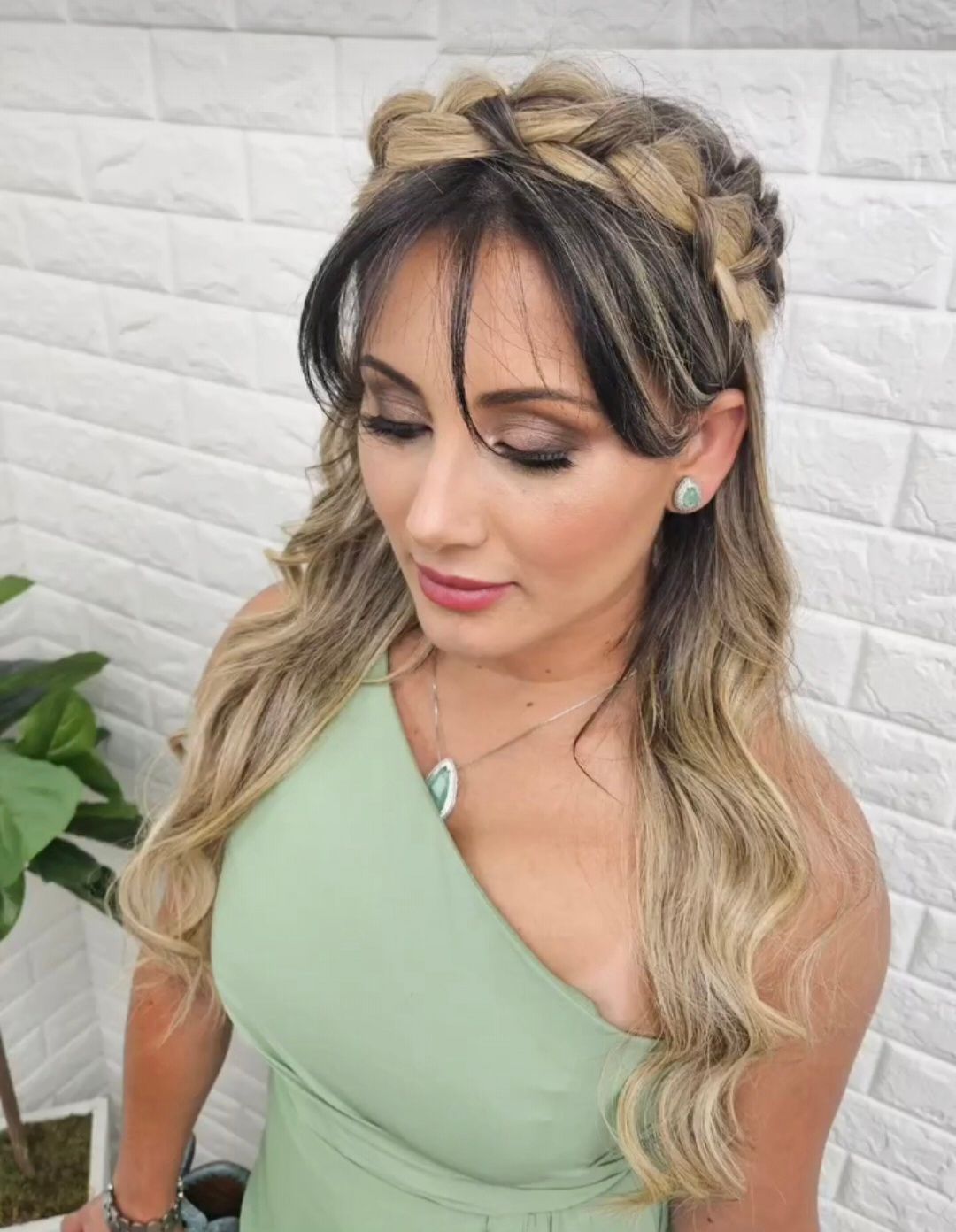 Woman with a braided hair crown, wearing a green one-shoulder dress, with wavy blonde hair.