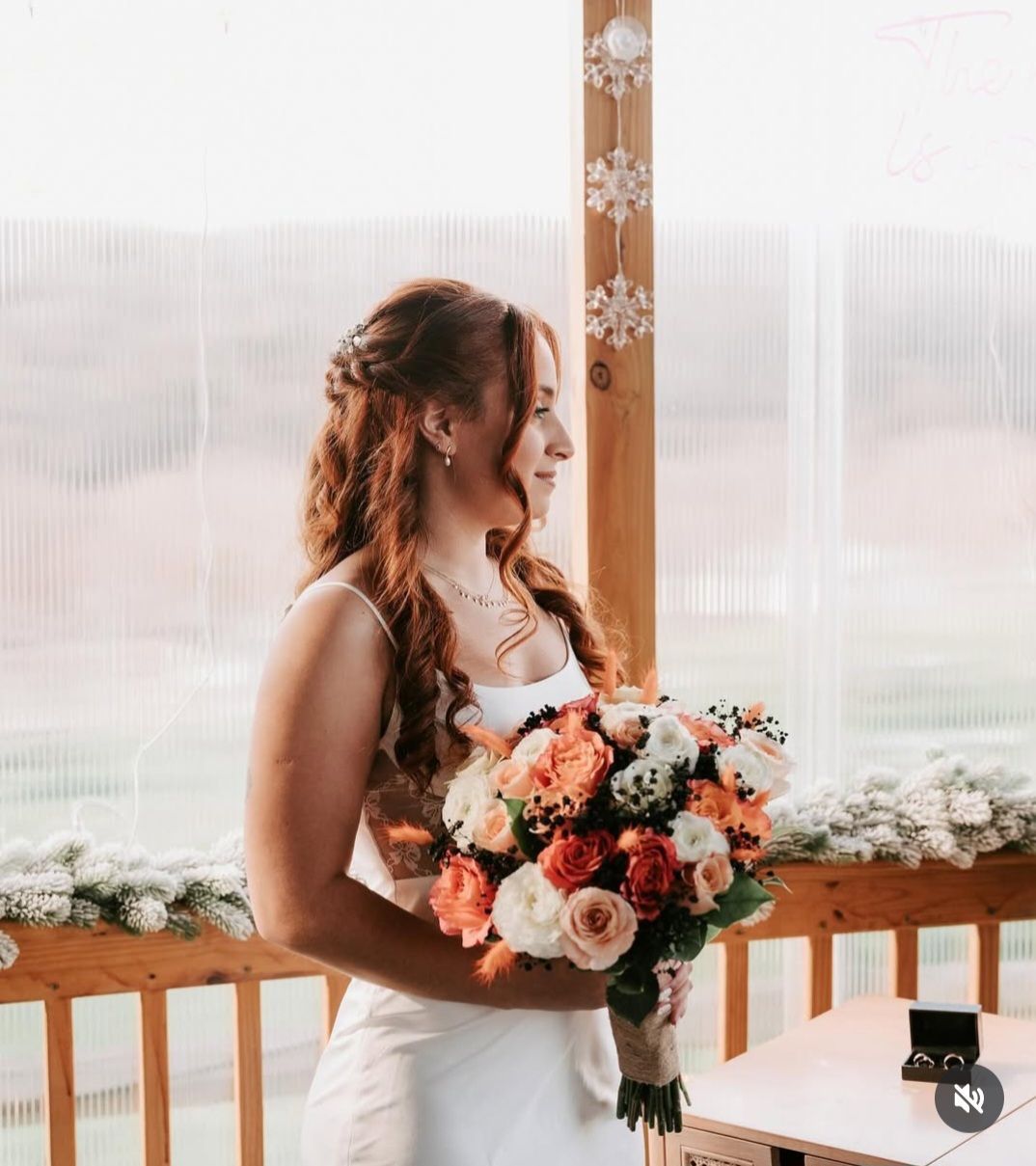 Bride in a white dress holds a bouquet, looking to the side; wooden railing and neutral background.