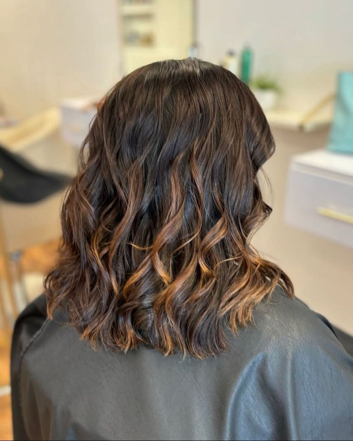 Woman's wavy, shoulder-length brown hair with caramel highlights. In salon setting, wearing black cape.