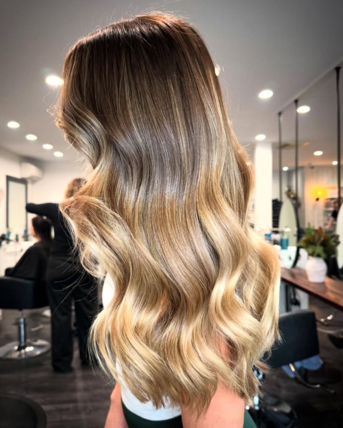 Woman with long wavy hair, brown at the roots, transitioning to blonde. Salon setting with mirrors.