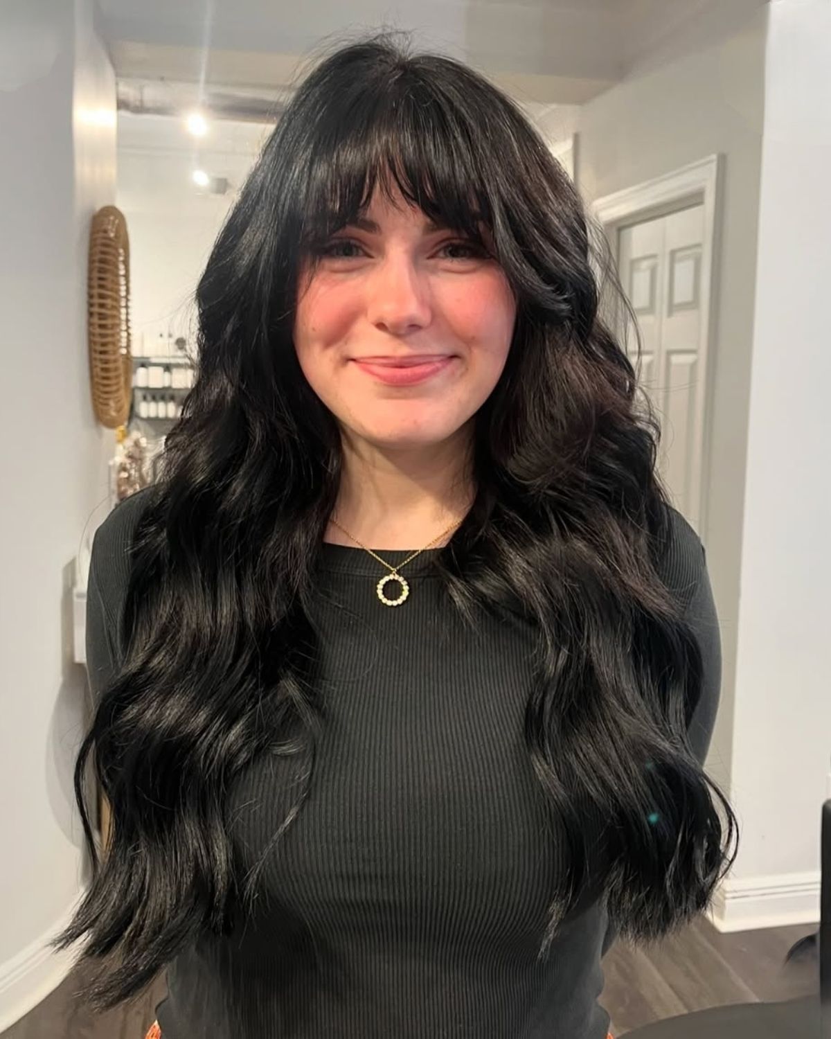 Woman with long, wavy black hair and bangs, wearing a black top. Standing indoors, smiling.