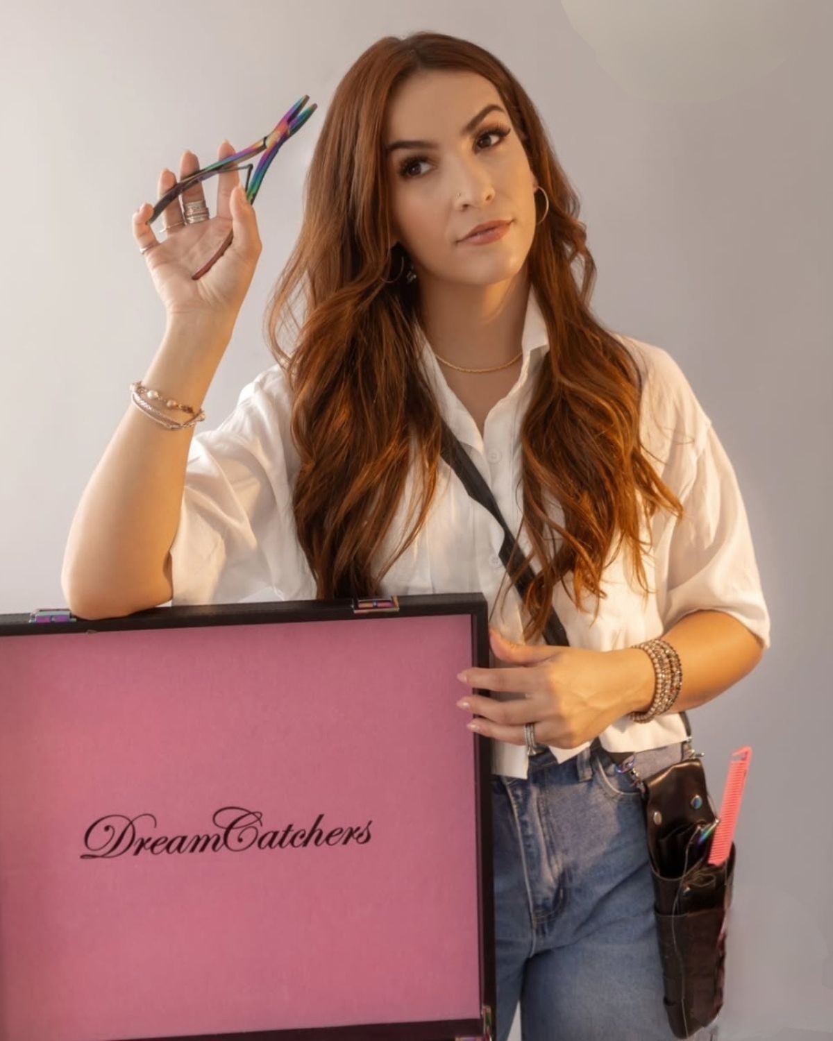 Woman holding hair tools, leaning on a pink DreamCatchers case. She wears a white shirt and jeans.