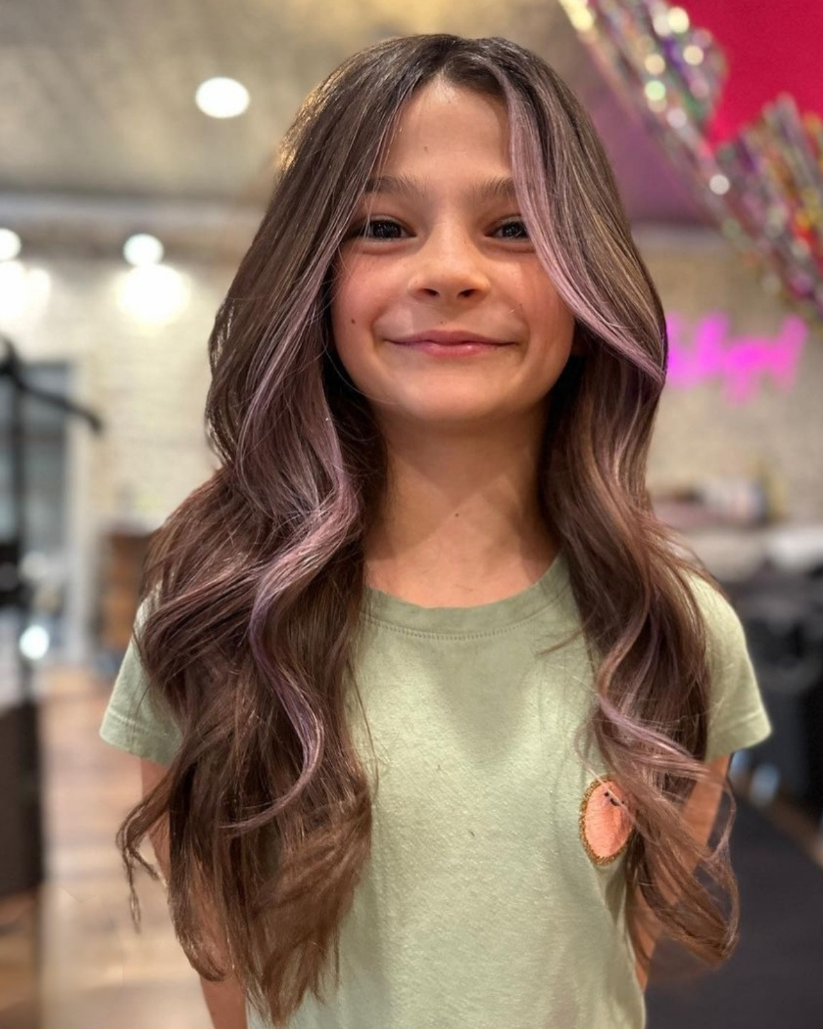 Girl with long wavy brown hair, pink highlights, smiling in a hair salon.