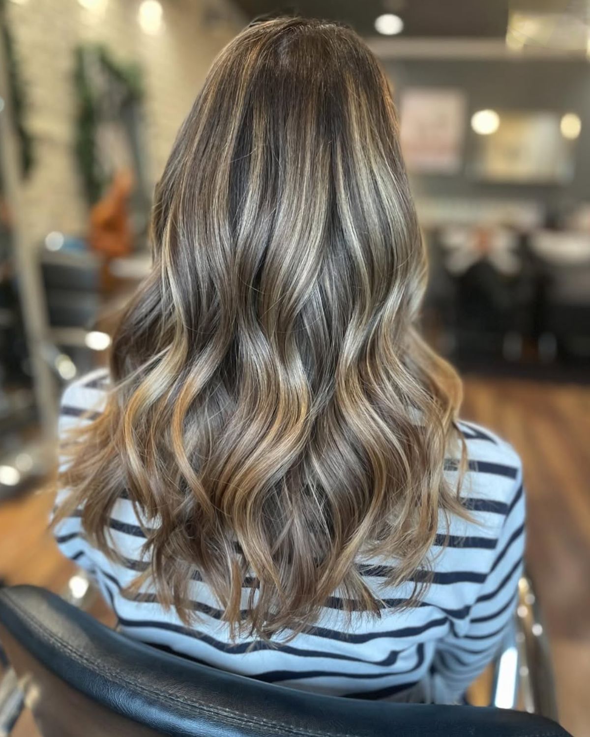 Woman with wavy hair; brown with blonde highlights, wearing a striped shirt. Sitting in a salon chair.