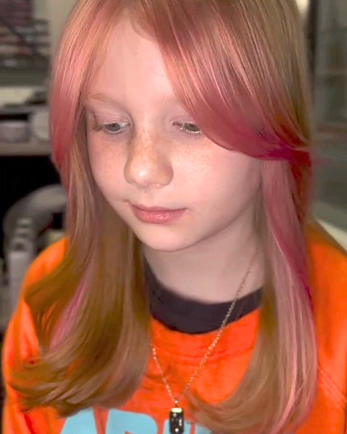 Girl with long hair, pink highlights, wearing orange shirt, looking down, indoor setting.