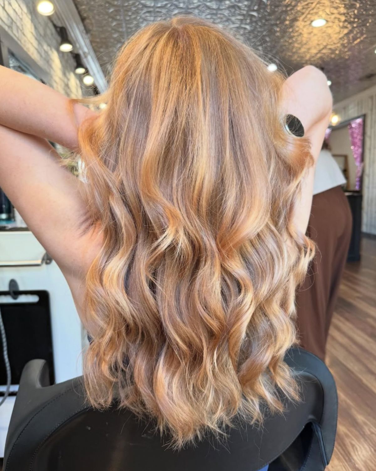 Woman with wavy, light blonde hair in a salon, hands behind head.