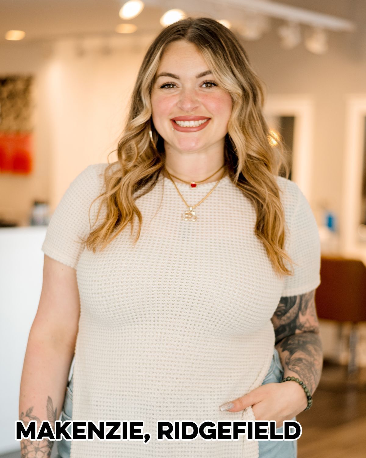 Woman smiling, standing in a salon, wearing a cream top and necklaces, tattoo on her arm.
