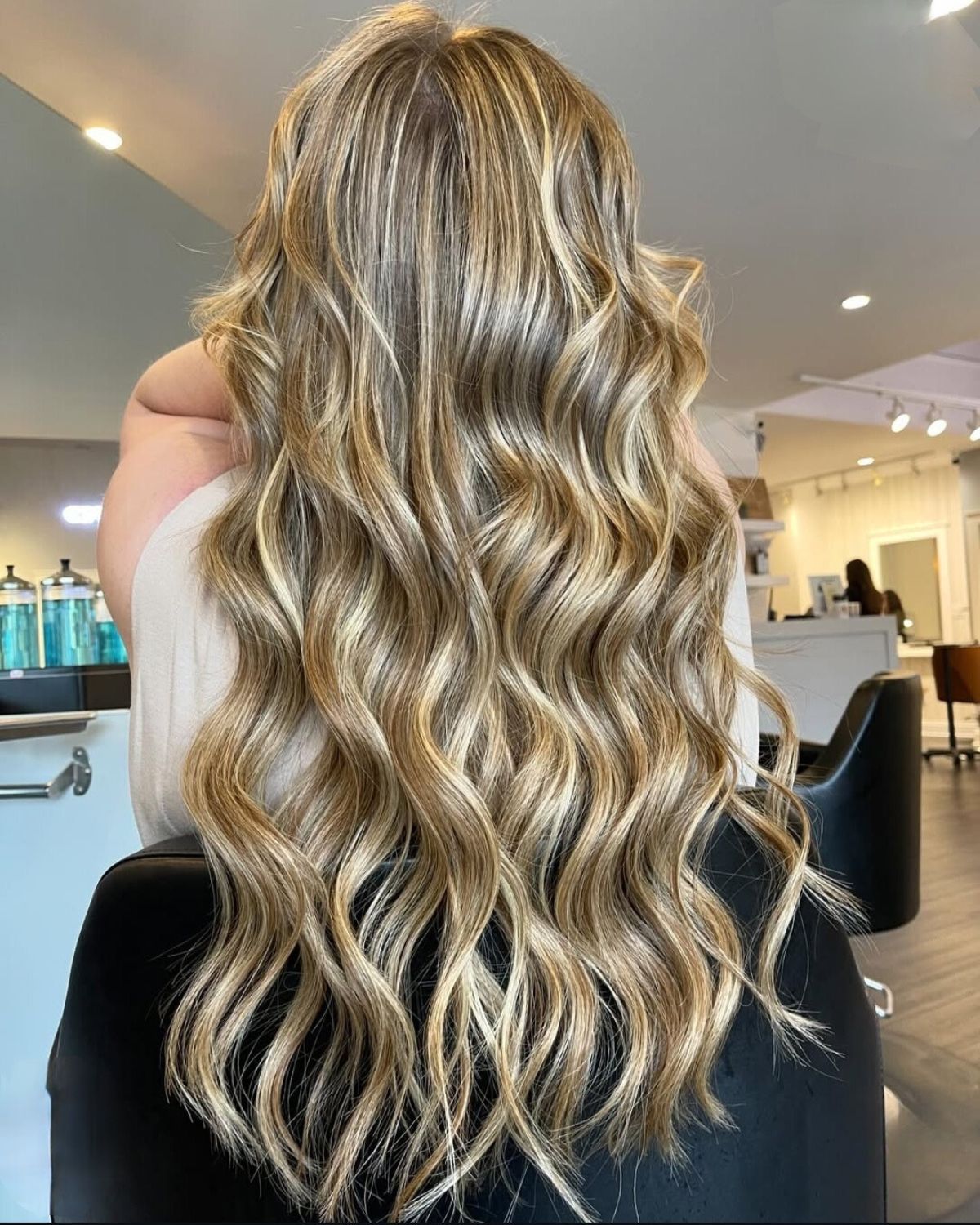 Woman with long, wavy blonde hair with highlights, sitting in a salon chair.