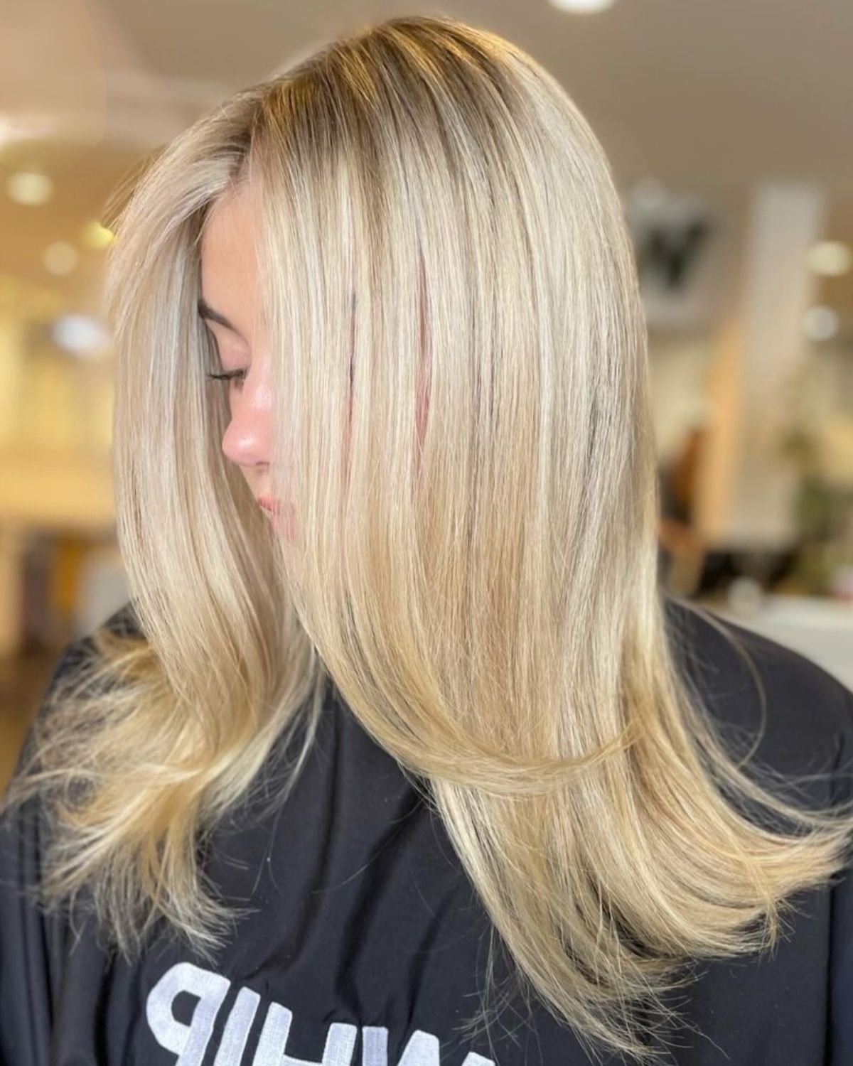 Blonde hair with highlights, styled straight with layers. Person facing partially left, wearing a black smock.
