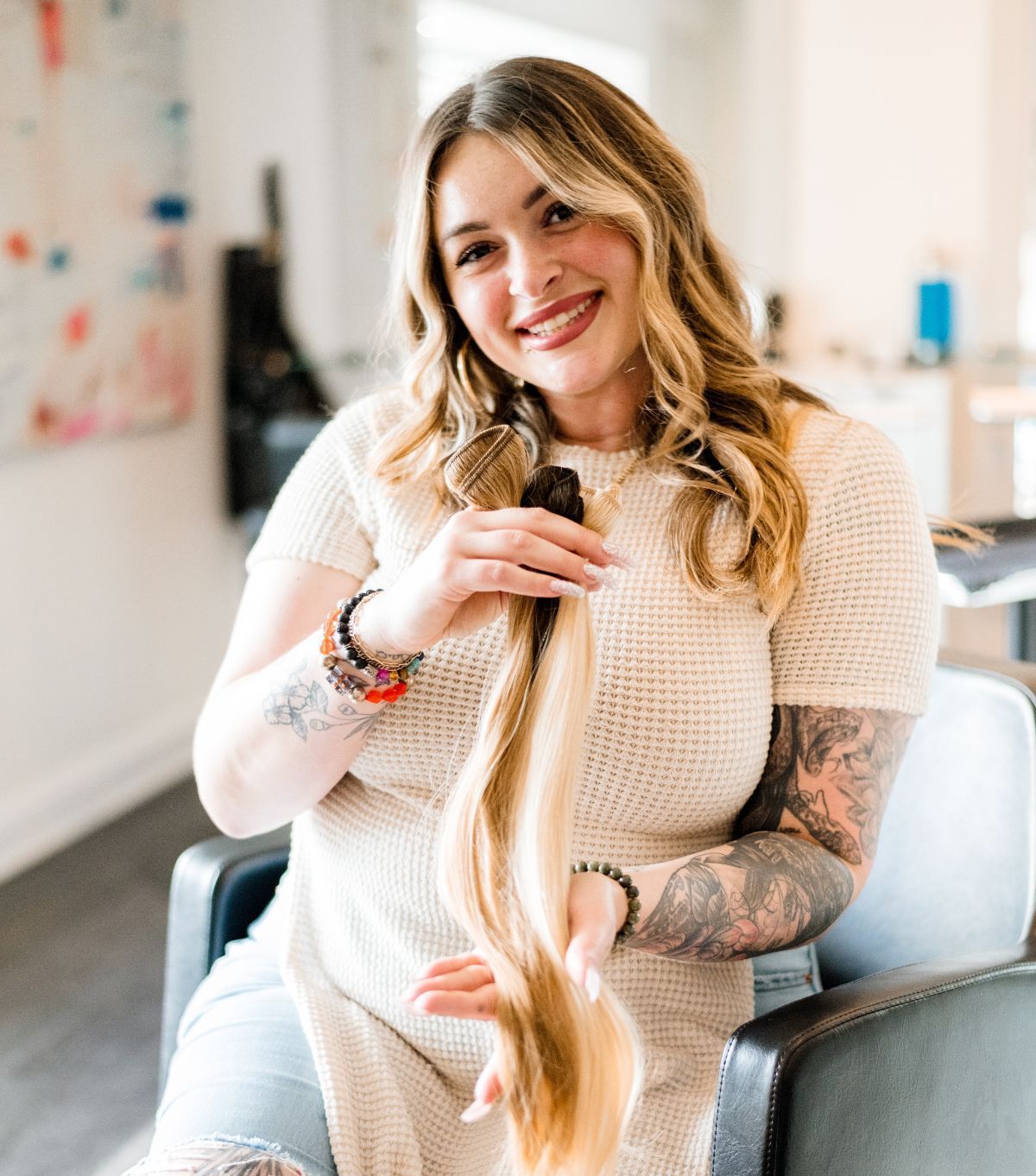 Woman smiles, holding a bundle of blonde hair extensions. She has tattoos and wears a cream-colored sweater.