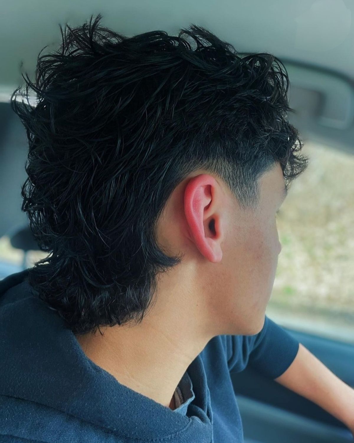 Person with dark, textured hair and a mullet hairstyle, looking right, in a car.