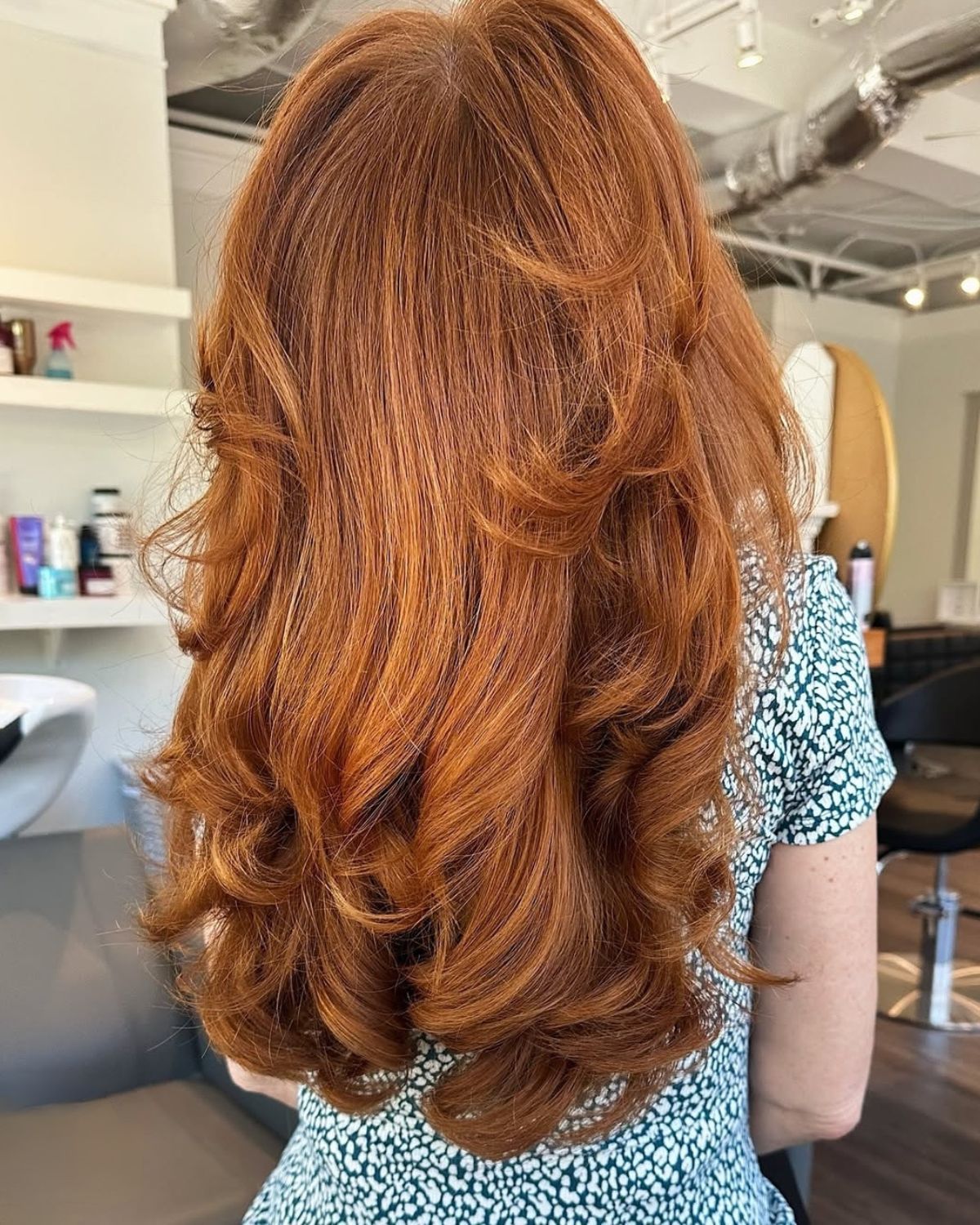 Woman with long, wavy auburn hair. She is wearing a teal and white floral patterned shirt. In a salon.
