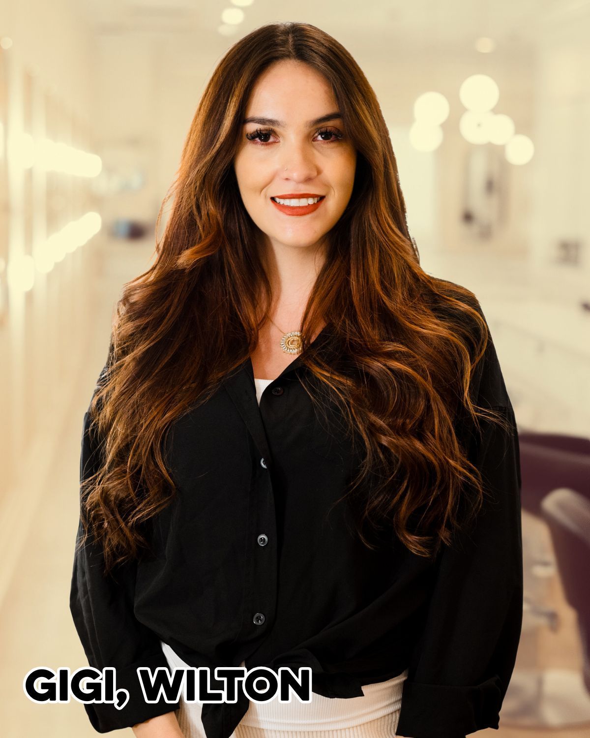 Woman with long, wavy brown hair, smiling, wearing a black shirt in a salon setting.