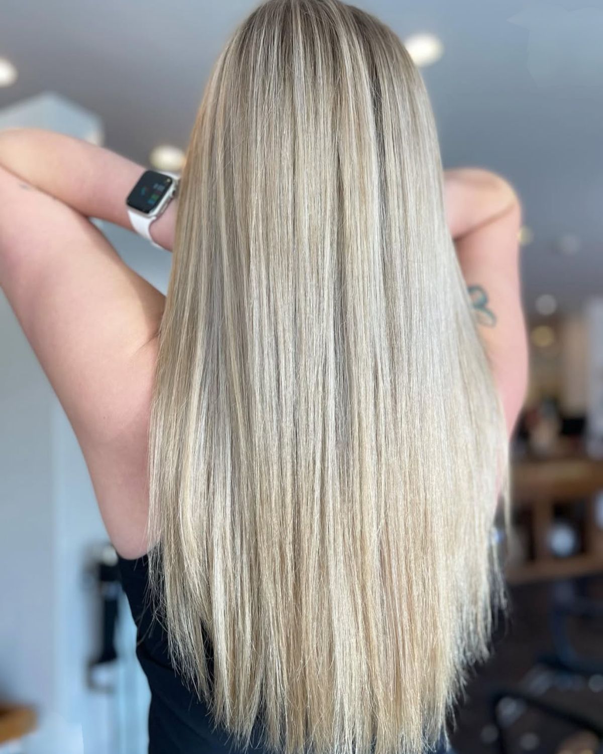Blond hair styled straight, reaching the waist; woman's back to camera, arms raised, wearing a watch.