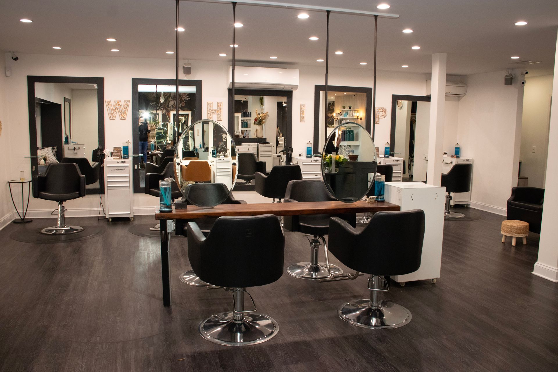 Hair salon interior with mirrors, chairs, and styling stations. Dark wood flooring, neutral walls, and overhead lighting.