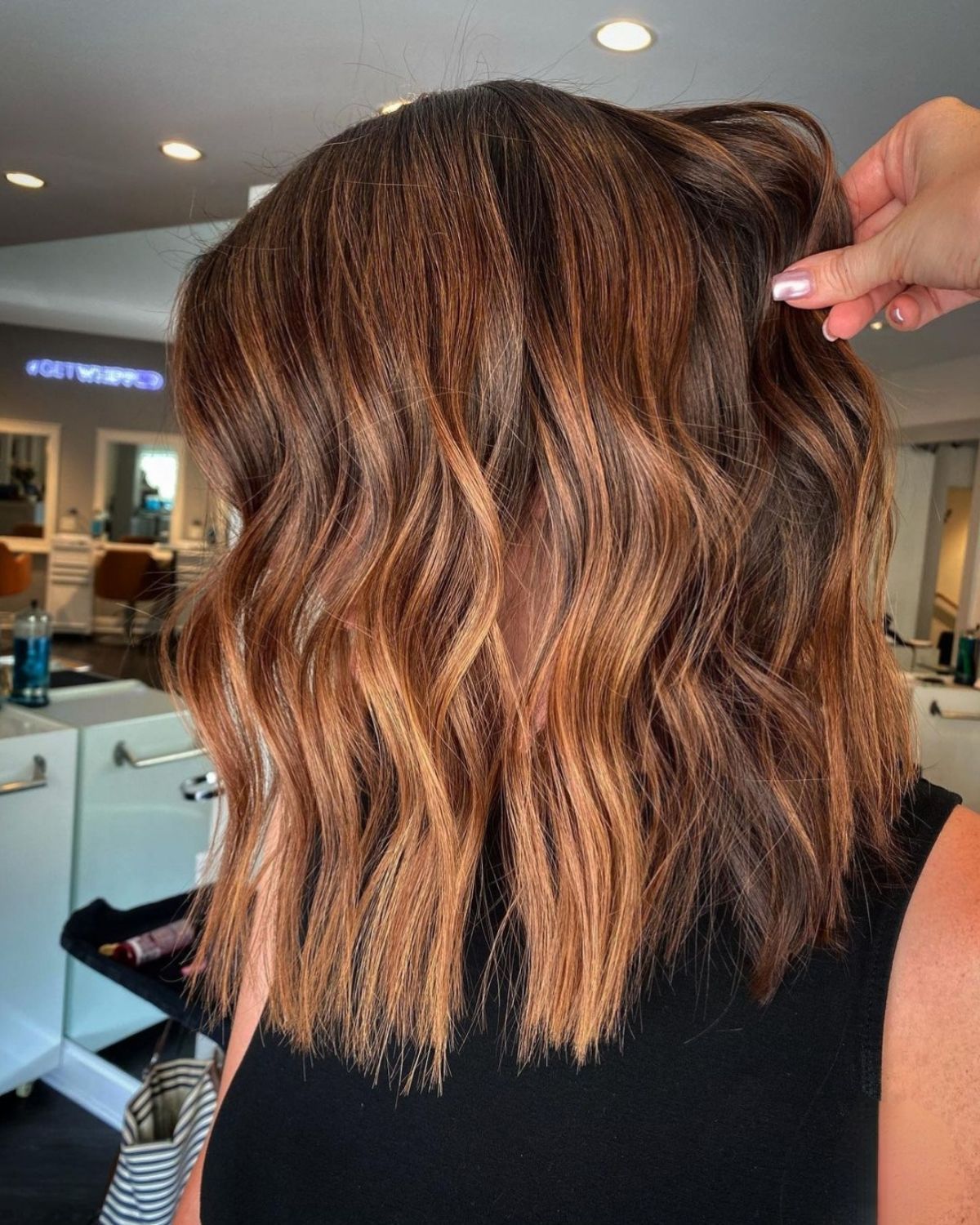 Brown hair with caramel highlights, styled in loose waves, in a salon. A hand touches the hair.