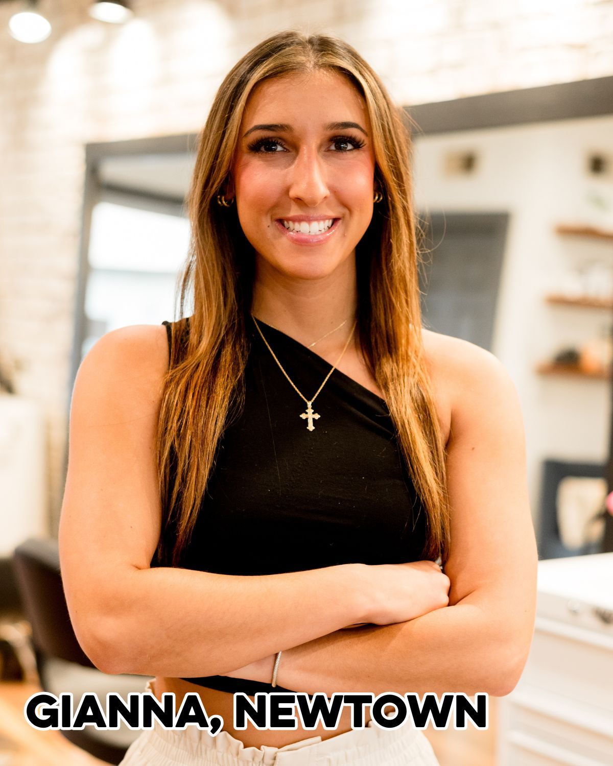 Woman, Gianna, with long brown hair smiles, arms crossed, in a salon, wearing black top.