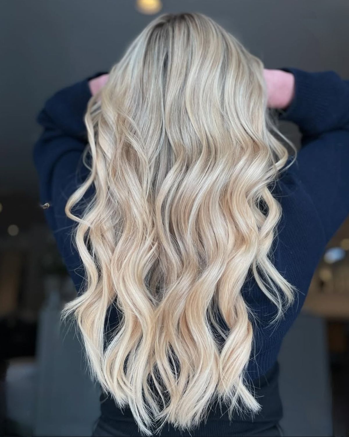 Blonde wavy hair cascading down back, hands near head, dark top.