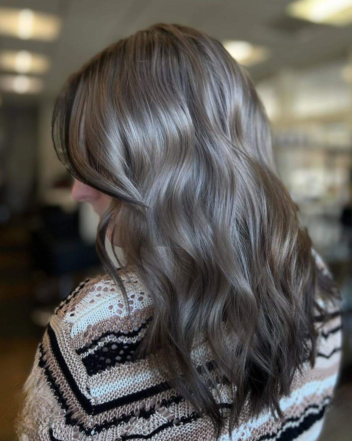 Woman with wavy, ash brown hair; wearing a patterned sweater, in a salon setting.