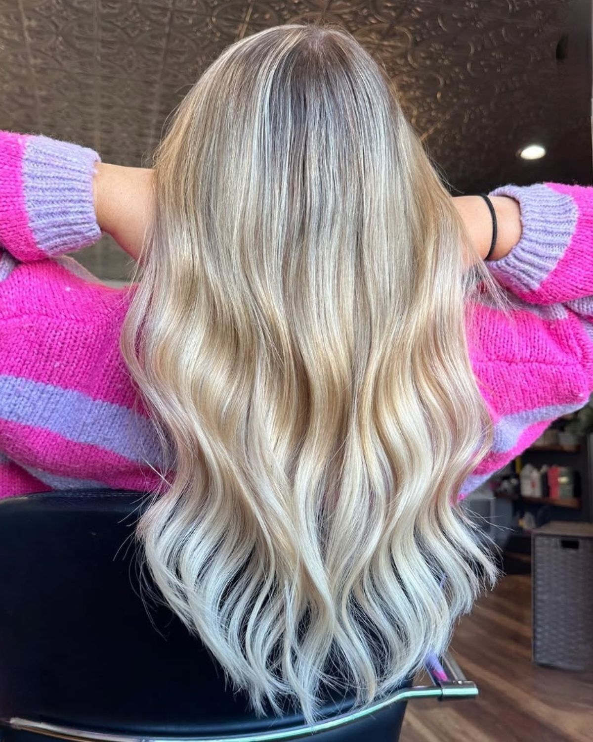 Woman with long blonde wavy hair, wearing a pink and purple sweater.