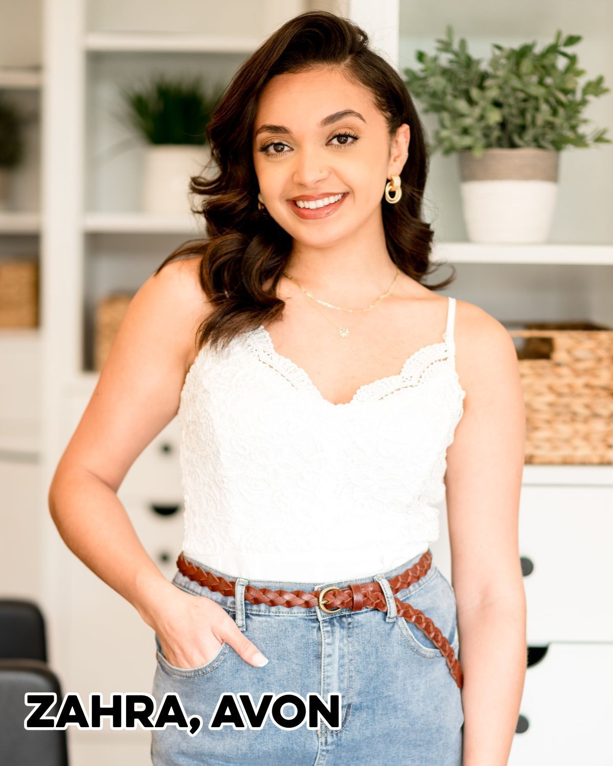 Woman in white top and jeans smiles, hands in pocket, brown belt. Shelf with plants in background.