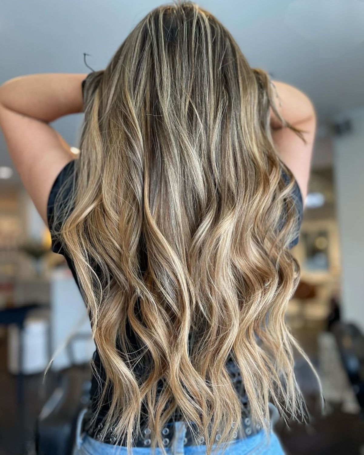 Woman with long, wavy, blonde and brown highlighted hair, arms raised, in a salon setting.