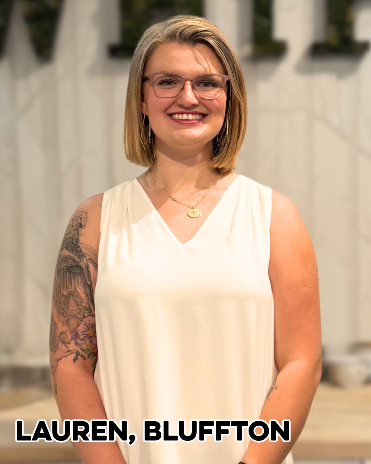 Woman, Lauren, with blonde bob, glasses, and arm tattoo smiles in front of a building.