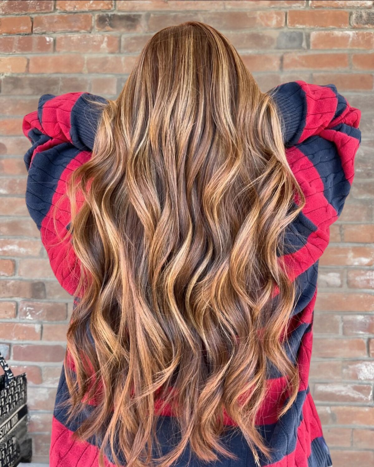 Long wavy brown hair with blonde highlights against a brick wall. Person wearing a red and navy sweater.