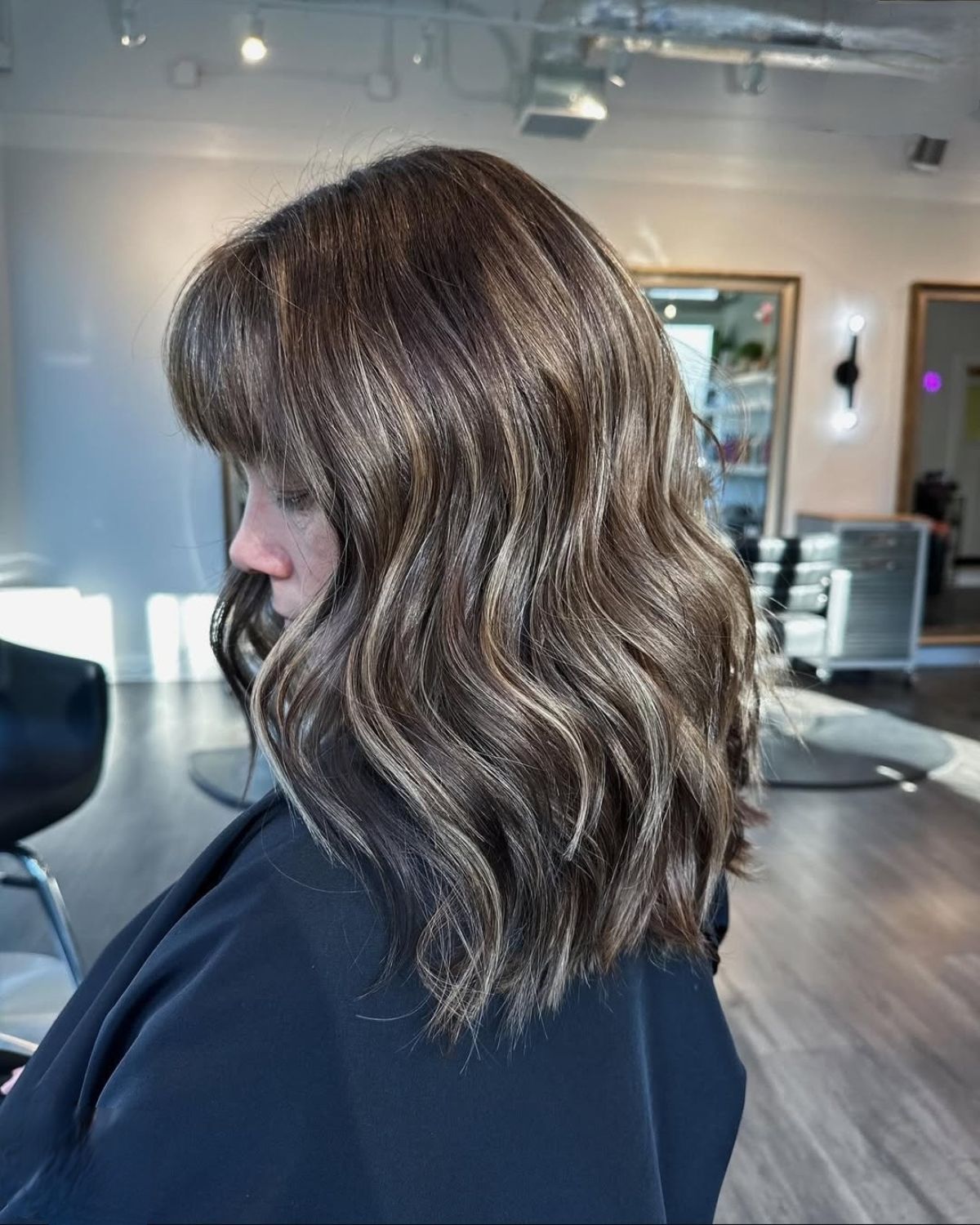 Woman with brown hair, face turned sideways, styled with waves and highlights, in a hair salon.