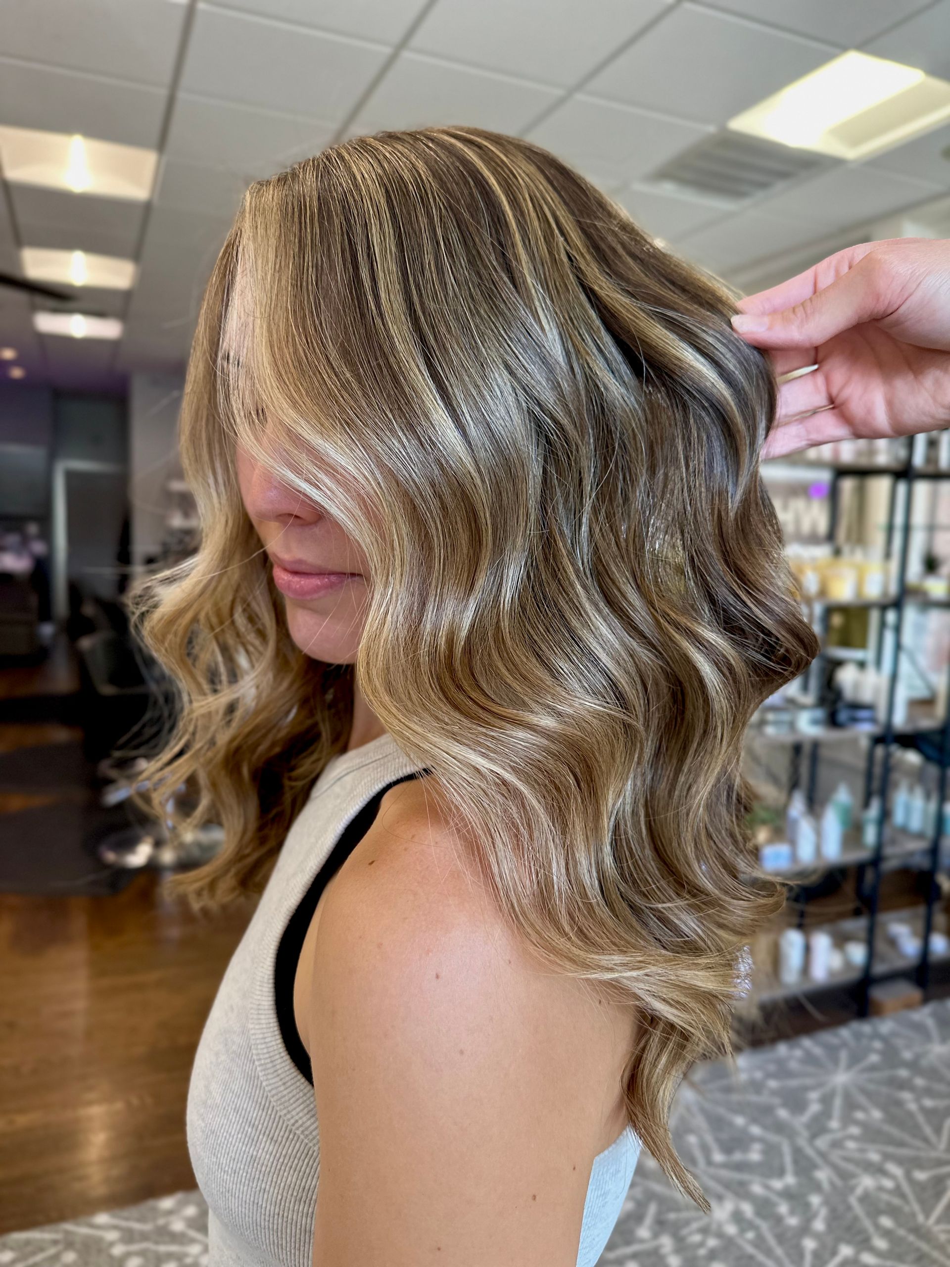 Woman with wavy highlighted hair, hand touching it. Indoors, salon setting.