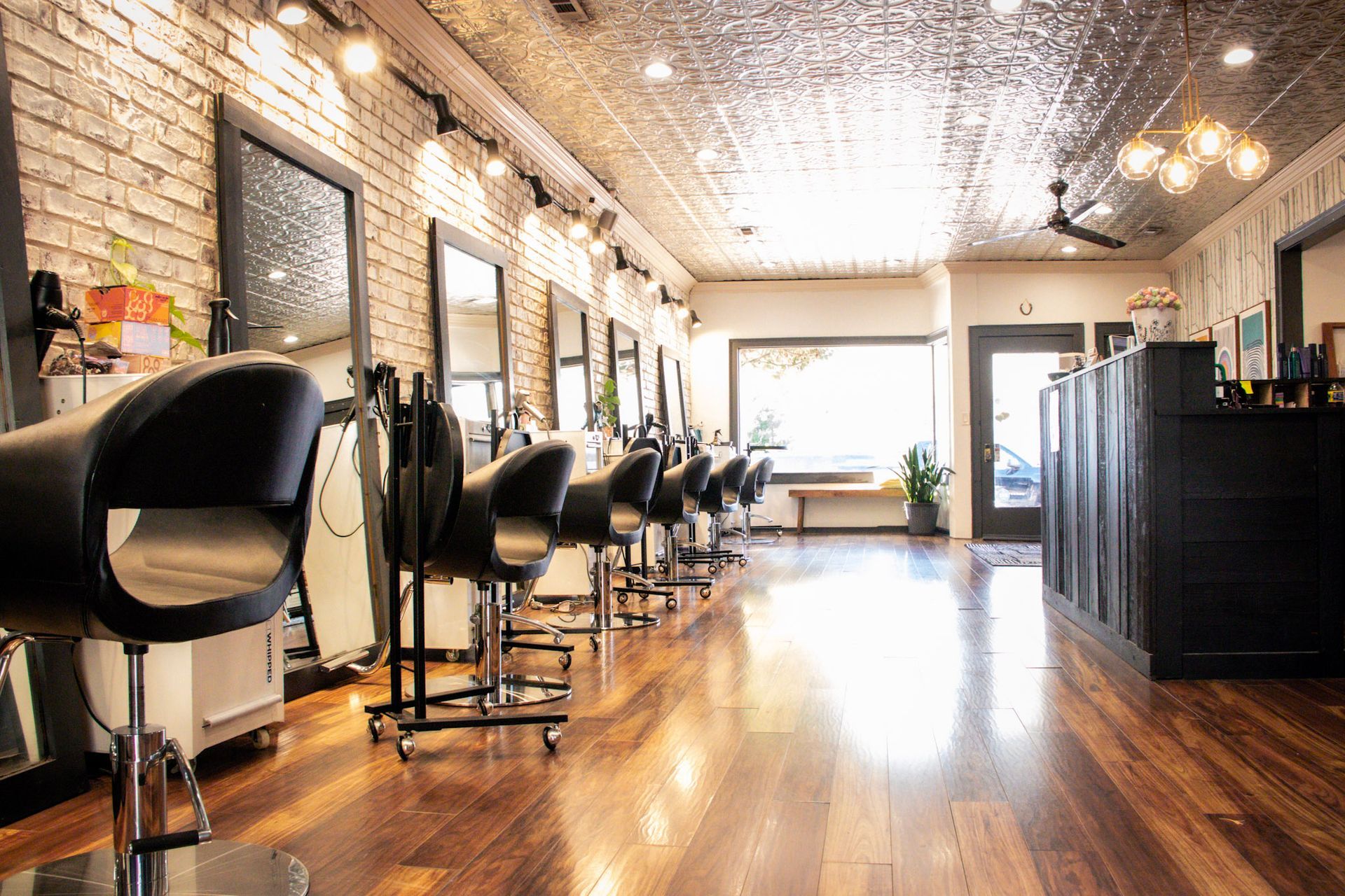 Hair salon interior: mirrors, chairs, brick wall, wood floor, and large window.