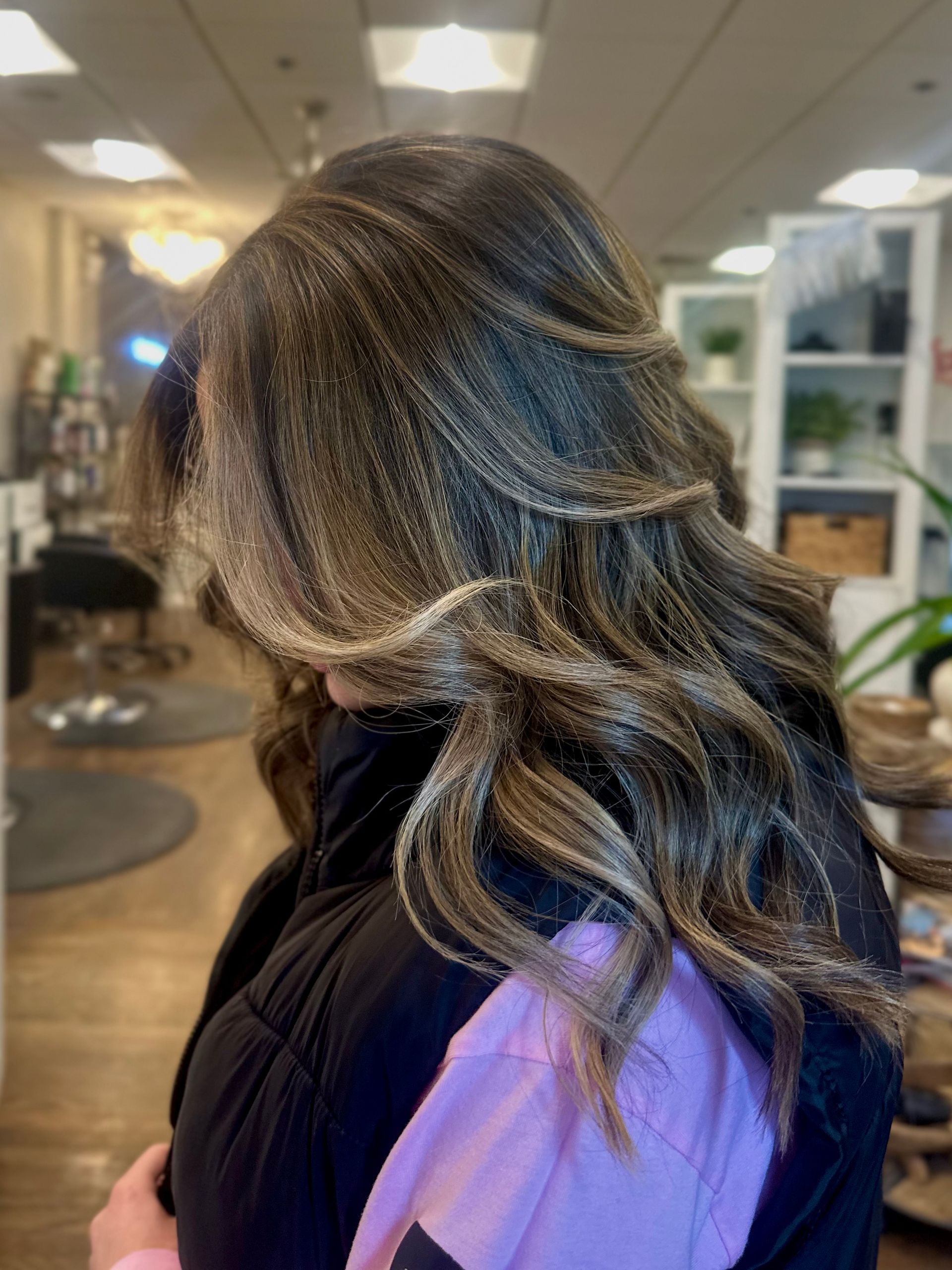 Woman with layered brown hair, highlighted with blonde tones, styled in waves. Indoors, blurred background.