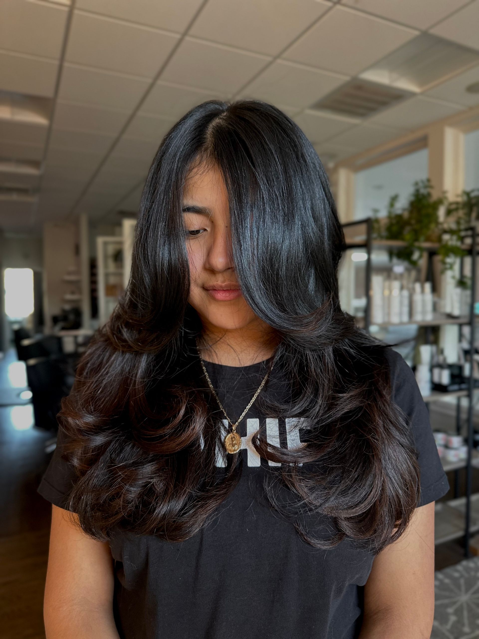 Woman with long, layered, dark hair in a salon; she's wearing a black shirt, looking down.