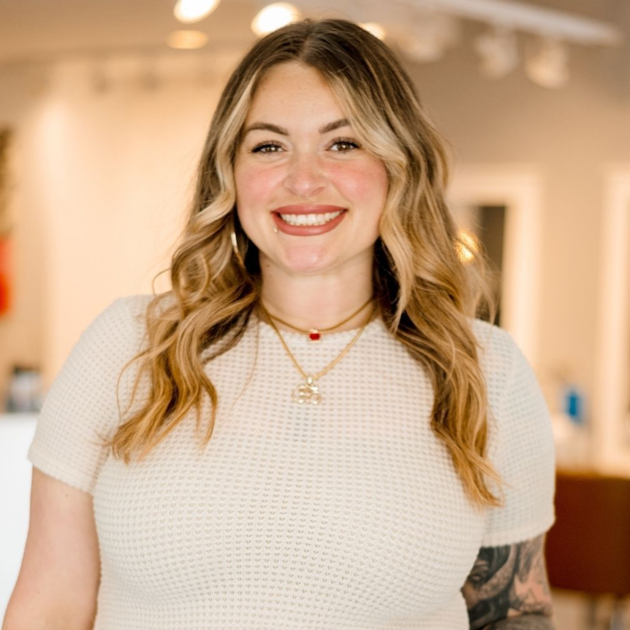 Woman with wavy blonde hair smiling, wearing a cream top and necklaces, in a brightly lit room.