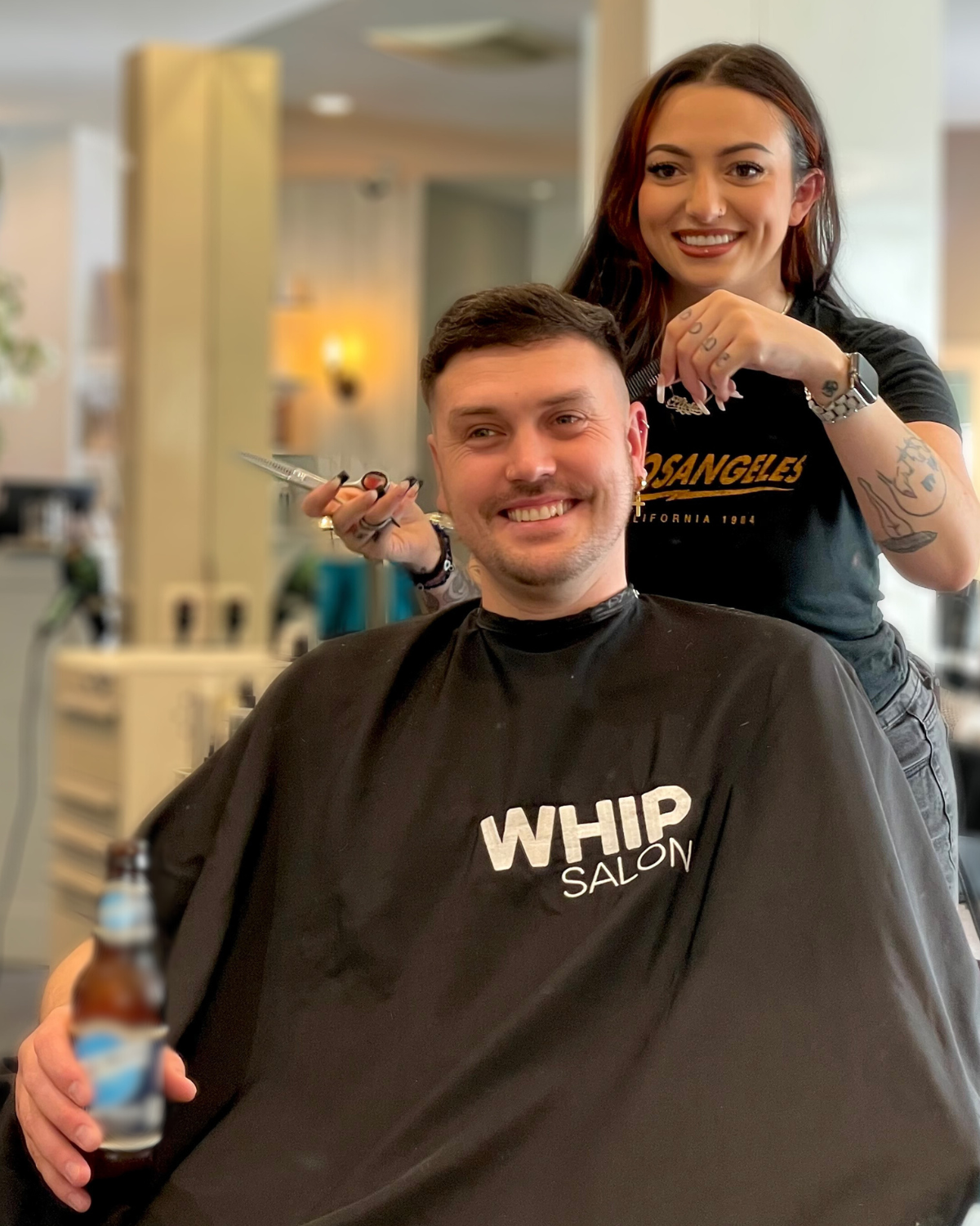 Man getting a haircut at a salon. Hairdresser smiles, holding scissors. Man smiles, wearing salon cape, holding beverage.