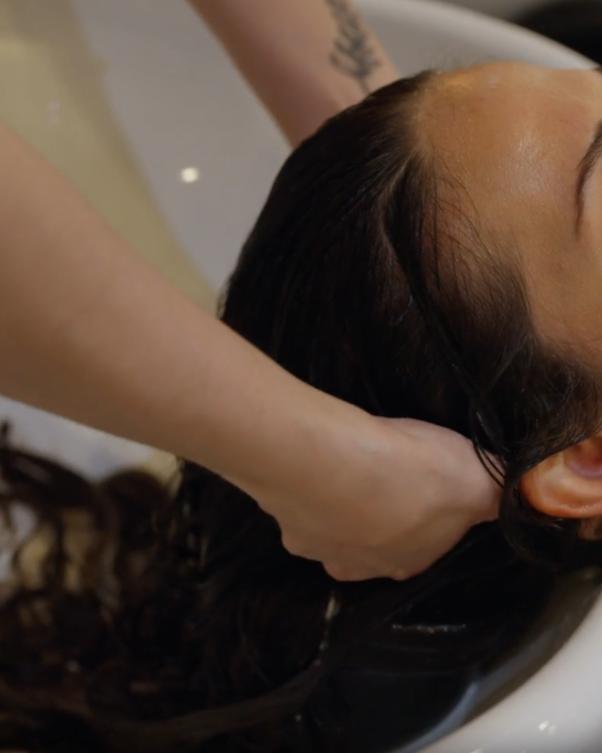 Person having their hair washed at a salon, head tilted back over a sink. A hand massages their hair.
