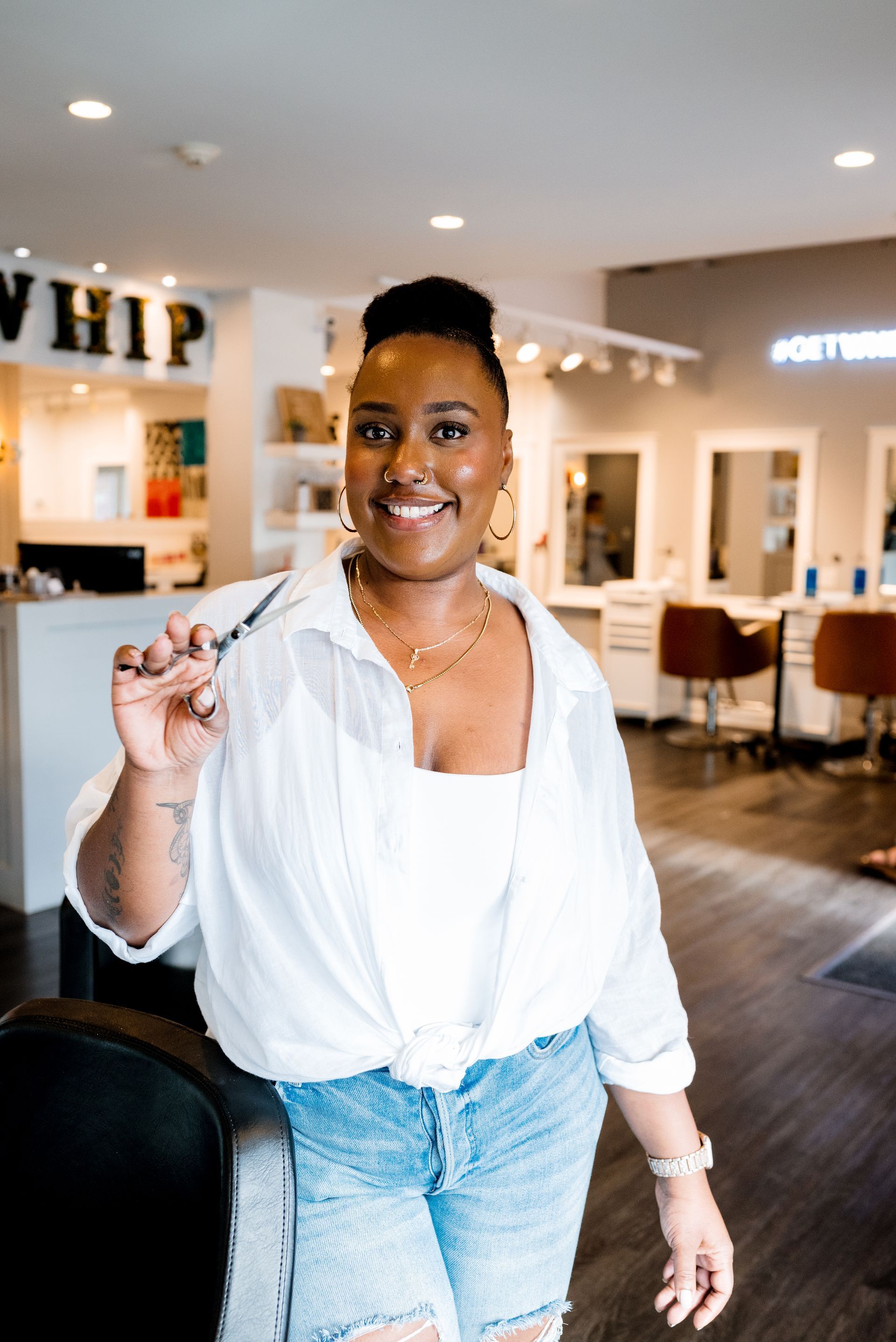 Smiling person in a salon, holding up scissors. Wearing a white shirt, jeans. The salon has brown and white decor.