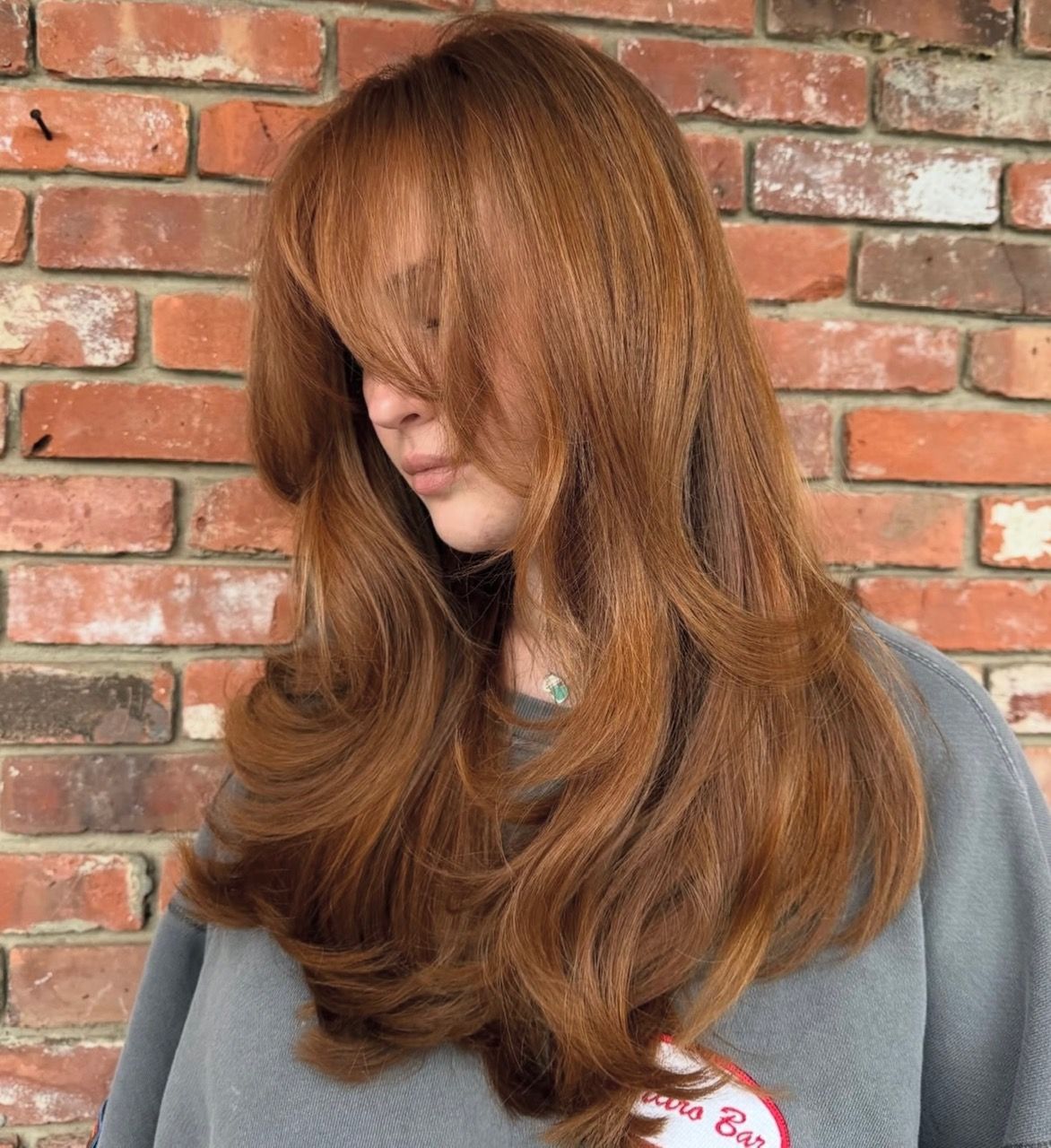 Woman with long, reddish-brown hair, layered cut, side-swept bangs; in front of a brick wall.