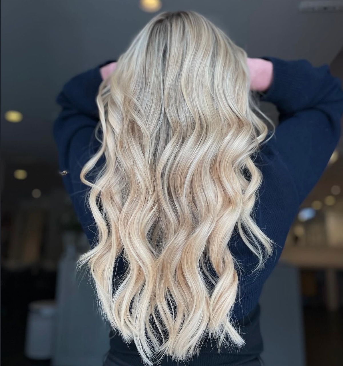 Blonde wavy hair styled down the back; arms raised. Indoors, neutral background.