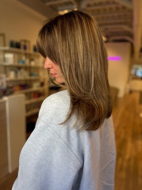 Woman with layered brown hair and highlights, wearing a light gray sweater.