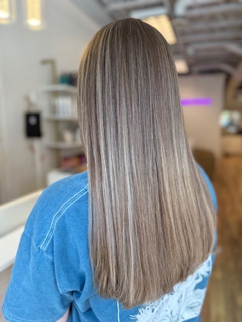 Woman with long, straight hair; brown with blonde highlights, wearing a blue shirt, in a salon.