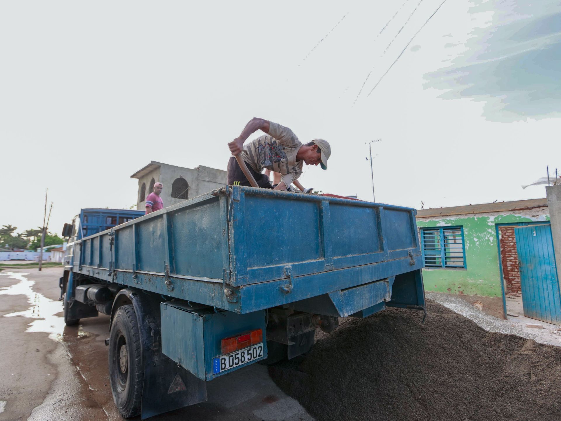 Worker shoveling gravel into a blue dump truck on a muddy road under overcast skies