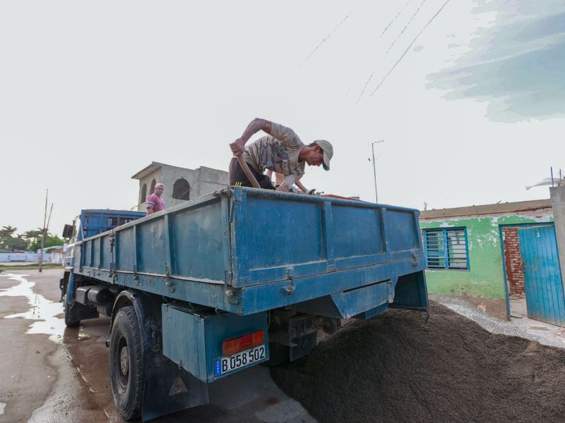 Worker shoveling gravel into a blue dump truck on a muddy road under overcast skies