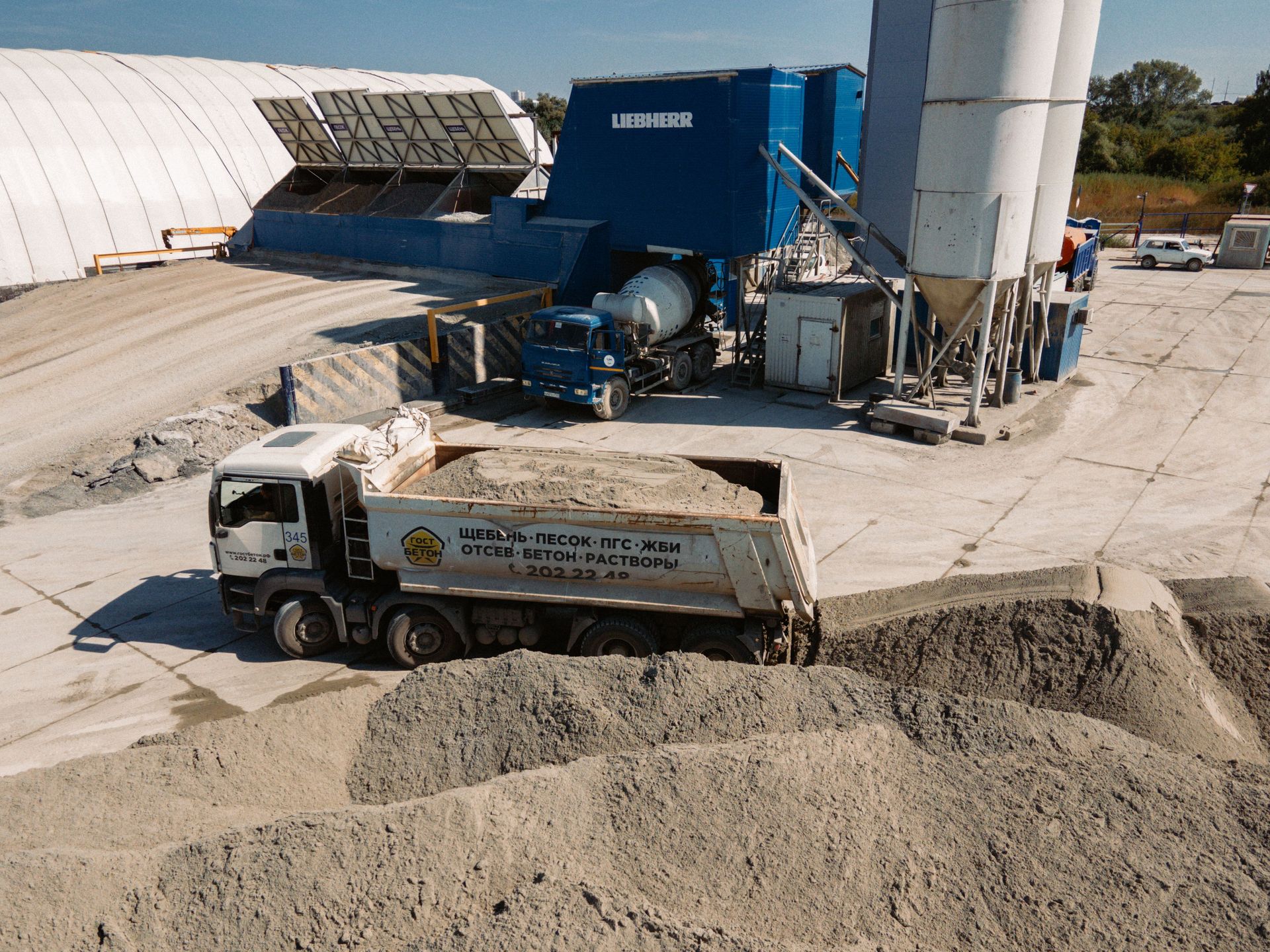 White truck dumps gravel at an industrial site beside a large blue machine and silos.