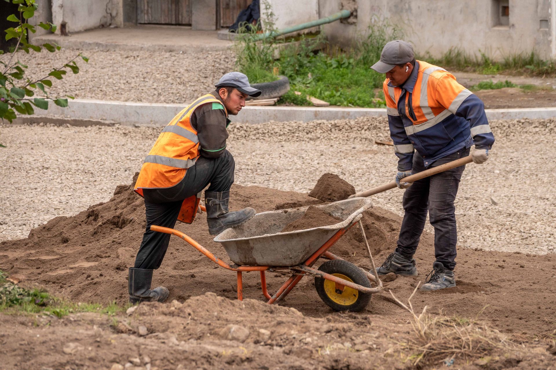 Two workers in orange vests pushing a wheelbarrow of dirt at a construction site