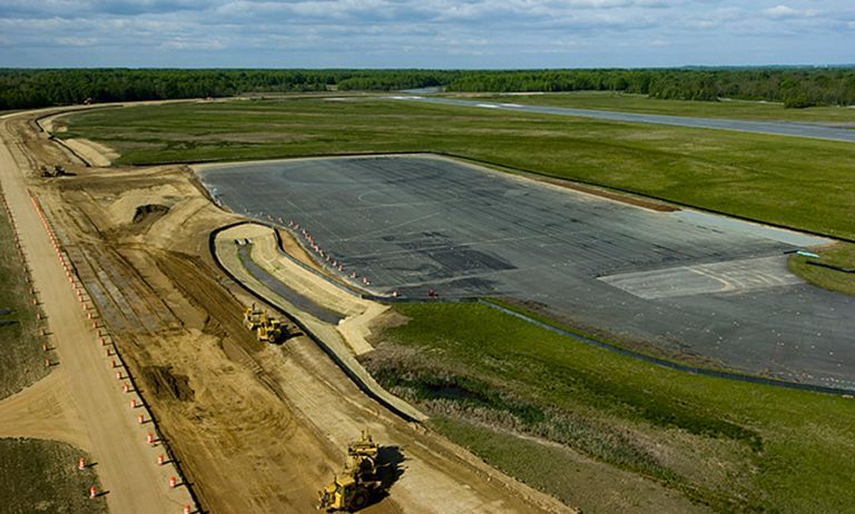 Aerial view of a construction site with a large lined pond beside green fields and a dirt embankment